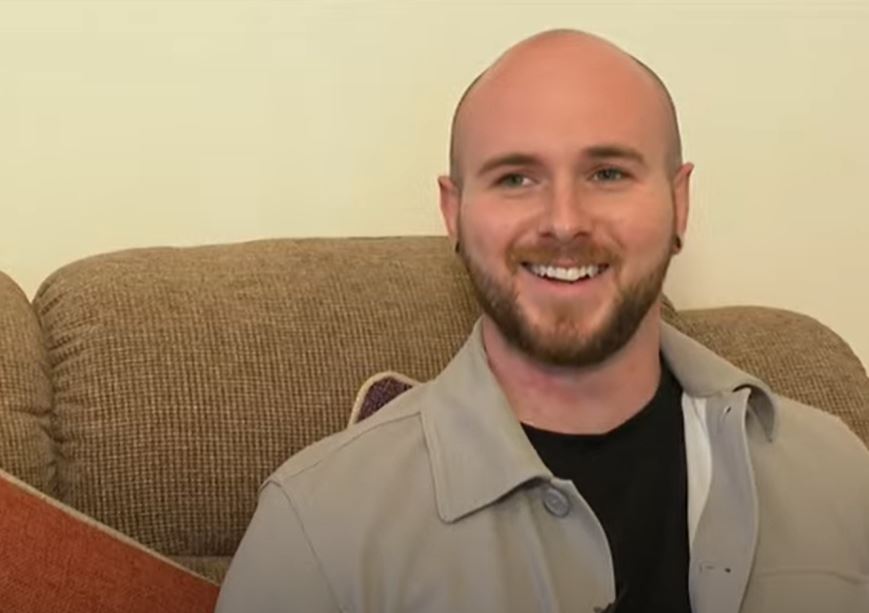 Luke, 27 form South Devon sits on a sofa. He is a smiling with very short hair and a little beard